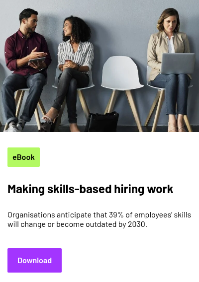 Three people seated on chairs against a wall; two are engaged in conversation while the third is focused on a laptop.